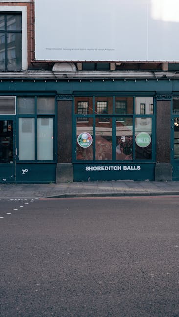 The image depicts a storefront with a dark teal-painted wooden frame and large window panels on the ground floor, positioned directly on a paved sidewalk in an urban setting. The windows feature small circular decals, with one displaying the words 'SHOREDITCH BALLS' in white lettering on a dark background, serving as the shop's sign. Above the storefront, part of an upper building structure is visible, including an exterior wall with a combination of white and exposed brick sections, indicating a mixed architectural style. The pavement in front appears clean and well-maintained, with no visible debris or obstructions, suggestive of a typical commercial street environment. The scene is illuminated by natural daylight, giving a neutral tone to the overall appearance. This type of setting might relate to independent waste handling or on-site clearance processes, often associated with local businesses or private property management services like Waste Clearance Shoreditch, supporting the context of rubbish removal or alternative waste disposal options in urban environments.