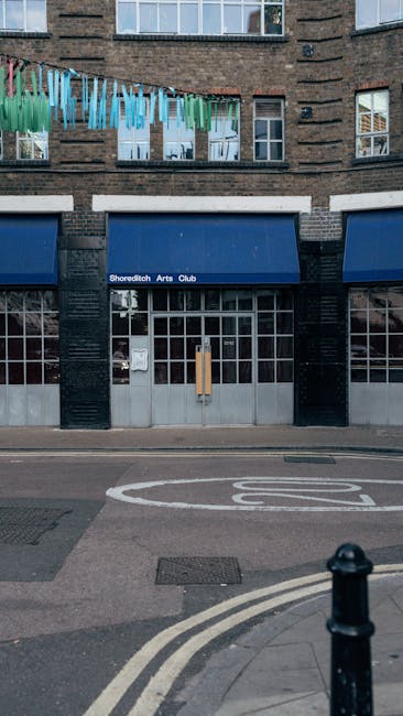 The image depicts a storefront with a dark teal-painted wooden frame and large window panels on the ground floor, positioned directly on a paved sidewalk in an urban setting. The windows feature small circular decals, with one displaying the words 'SHOREDITCH BALLS' in white lettering on a dark background, serving as the shop's sign. Above the storefront, part of an upper building structure is visible, including an exterior wall with a combination of white and exposed brick sections, indicating a mixed architectural style. The pavement in front appears clean and well-maintained, with no visible debris or obstructions, suggestive of a typical commercial street environment. The scene is illuminated by natural daylight, giving a neutral tone to the overall appearance. This type of setting might relate to independent waste handling or on-site clearance processes, often associated with local businesses or private property management services like Waste Clearance Shoreditch, supporting the context of rubbish removal or alternative waste disposal options in urban environments.
