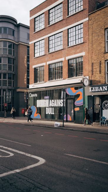 The image depicts the street-level view of a multi-storey brick building with large, grid-patterned windows in white frames. The ground floor features a glass storefront with a black background, displaying a large graphic with the text 'FASHION AGAINST DULL' and abstract, colorful shapes. Adjacent to the storefront, on the right, part of a black sign with white lettering is visible, indicating the presence of a nearby business. In front of the building, there is a paved sidewalk with pedestrians walking past; some are wearing casual clothing and carrying bags. The street has marked lanes and a curb, with a few small, scattered objects on the pavement near the pedestrians. The overall scene appears to be in an urban commercial district, with other modern buildings visible in the background, and the lighting suggests it is daytime with overcast skies. The setting aligns with urban environments where private waste clearance services like Waste Clearance Shoreditch might operate to support building renovations or business upgrades, highlighted indirectly by the absence of visible rubbish or clutter in the immediate scene.