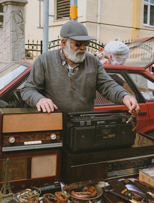 An elderly man with a grey beard, wearing a grey cap, sunglasses, and a grey sweater over a checked shirt, stands beside a red car with its window rolled down. He is holding a beaded necklace in one hand and is placing a portable black cassette player on top of a collection of vintage radios and electronic devices that are laid out on a table or stall. The scene appears to be outdoors, in a residential or urban area, with a stone building, a barred fence, and a yellow wall visible in the background. Various items including small figurines, jewelry, and miscellaneous objects are displayed on the table, indicating a market or private collection setting. The lighting is natural, suggesting daytime, and the overall scene reflects a typical second-hand sale or rubbish clearance where old electronic items and collectibles are being sorted or sold, exemplifying alternative waste handling or private disposal practices.