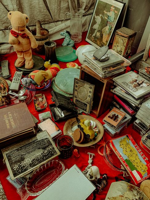 An elderly man with a grey beard, wearing a grey cap, sunglasses, and a grey sweater over a checked shirt, stands beside a red car with its window rolled down. He is holding a beaded necklace in one hand and is placing a portable black cassette player on top of a collection of vintage radios and electronic devices that are laid out on a table or stall. The scene appears to be outdoors, in a residential or urban area, with a stone building, a barred fence, and a yellow wall visible in the background. Various items including small figurines, jewelry, and miscellaneous objects are displayed on the table, indicating a market or private collection setting. The lighting is natural, suggesting daytime, and the overall scene reflects a typical second-hand sale or rubbish clearance where old electronic items and collectibles are being sorted or sold, exemplifying alternative waste handling or private disposal practices.